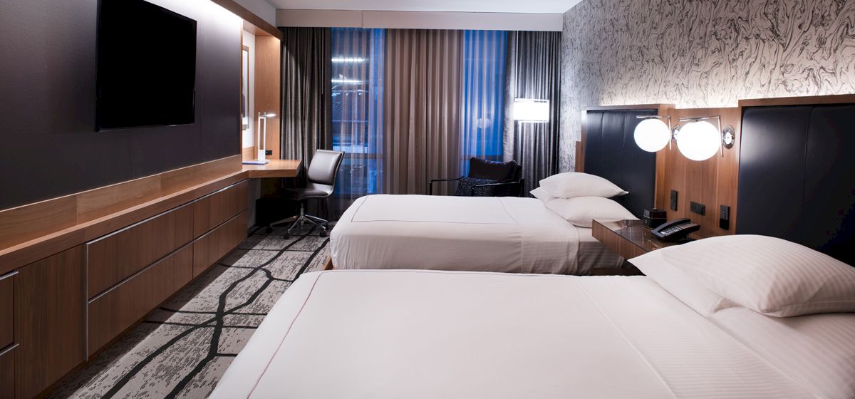The image shows a modern hotel room with two neatly made beds, a TV, desk, chair, and stylish lighting.