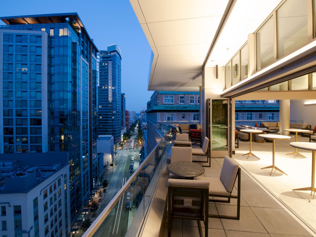 A modern cityscape at dusk with tall buildings, a well-lit balcony with seating, and a view of a street below.