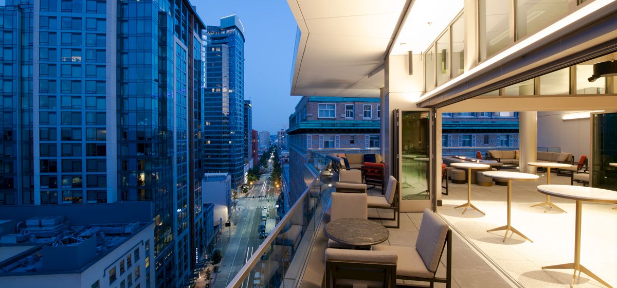 A modern cityscape at dusk with tall buildings, a well-lit balcony with seating, and a view of a street below.