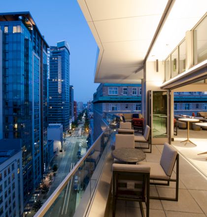 A modern balcony with furniture overlooks a city street lined with tall buildings at dusk.