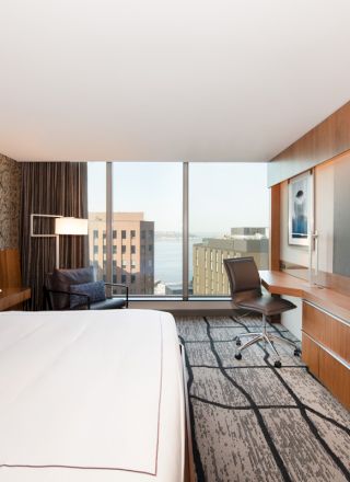 A modern hotel room with a large bed, desk, chair, and city view through a window. A TV is mounted on the wooden wall opposite the bed.