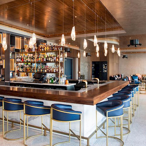 The image shows a modern bar with a sleek counter, blue barstools, hanging lights, and a well-stocked bar. Cozy seating is in the background.