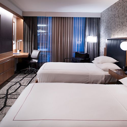 A modern hotel room with two beds, a TV, a desk, and ambient lighting, featuring a patterned carpet and wall.