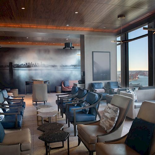 The image shows a modern lounge area with comfortable seating, large windows offering a scenic view of the waterfront and a city mural.