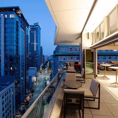 A modern balcony view overlooking a city street with tall buildings, featuring outdoor seating and warm lighting from the building interior.