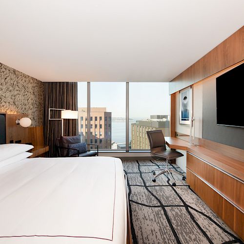 A modern hotel room with a large bed, flat-screen TV, desk, chair, and window view of buildings and water outside.