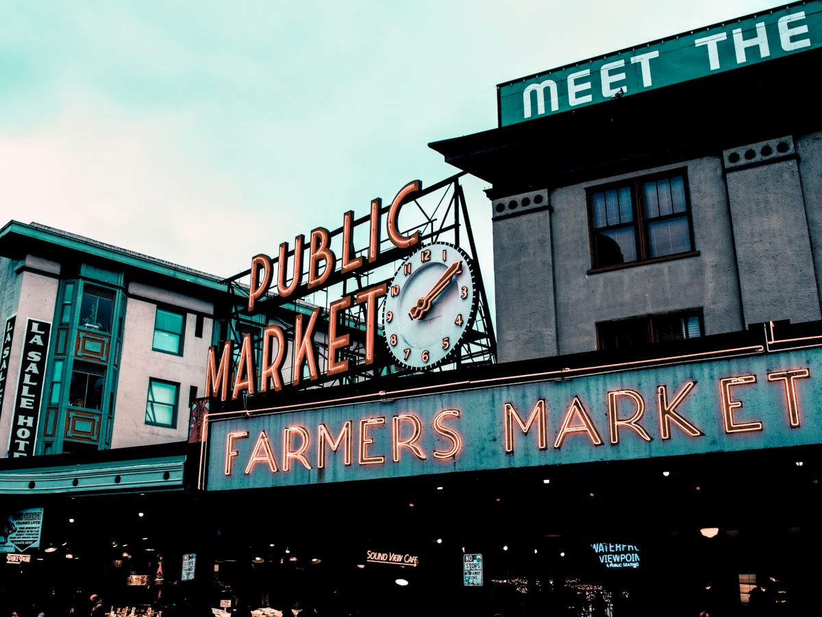 The image features a neon sign for a "Public Market" and "Farmers Market" with a clock, set against an urban building backdrop.