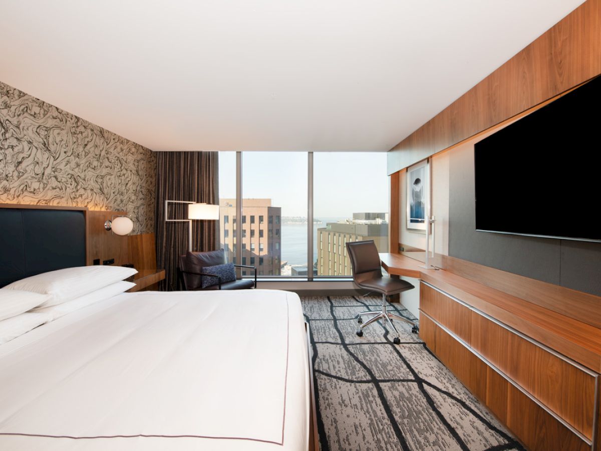 A modern hotel room features a large bed, flat-screen TV, sleek desk, and city view through a window, with stylish decor.
