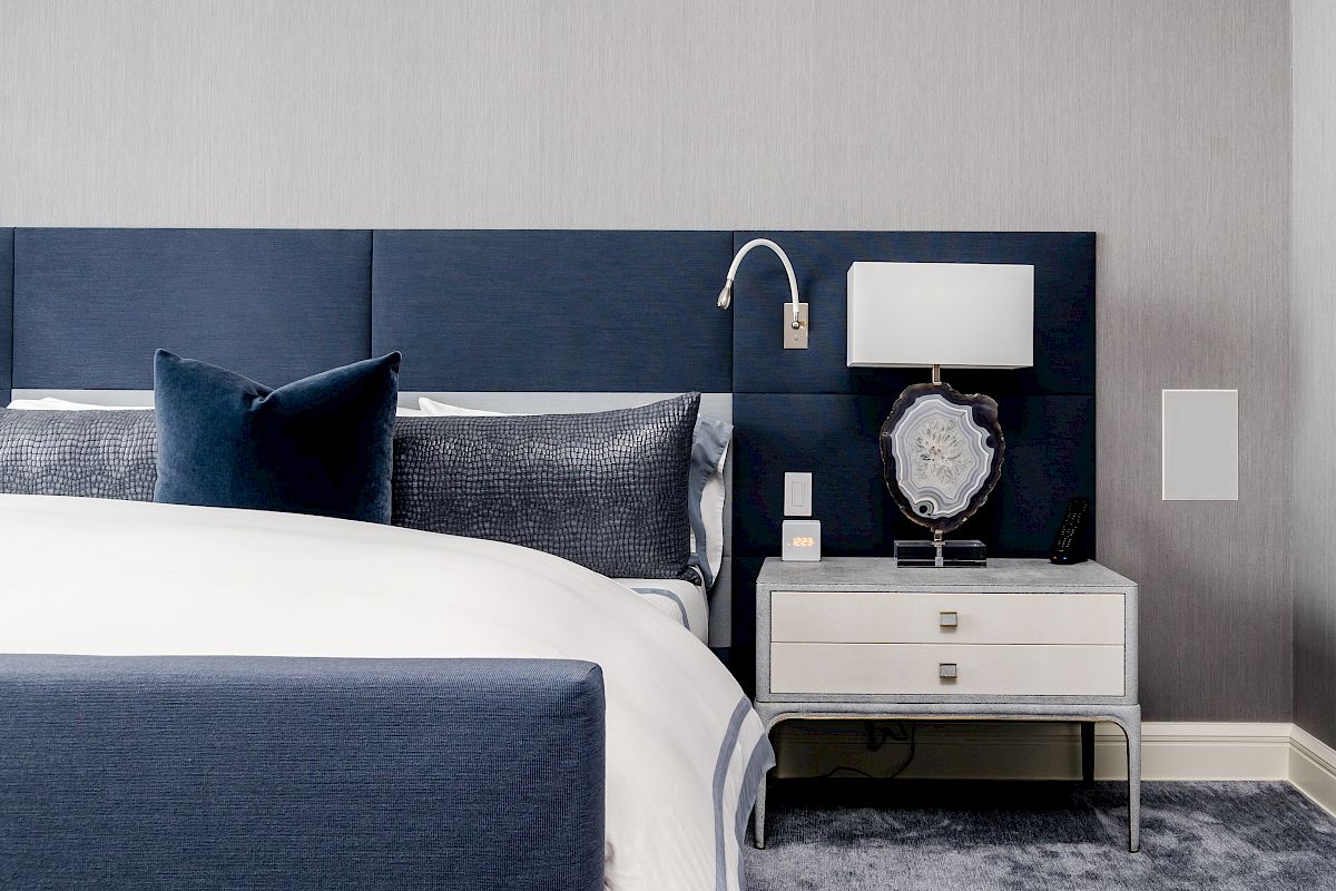 A modern bedroom with a navy headboard, plush white bedding, a nightstand with a lamp, and a gray carpeted floor completes the look.