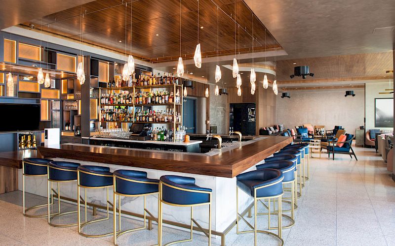 A chic bar with blue stools, hanging lights, and a well-stocked liquor shelf; modern seating area in the background.