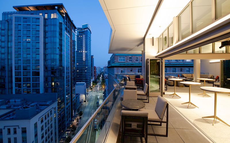 A modern balcony with seating overlooks a city street lined with tall buildings, under a twilight sky.