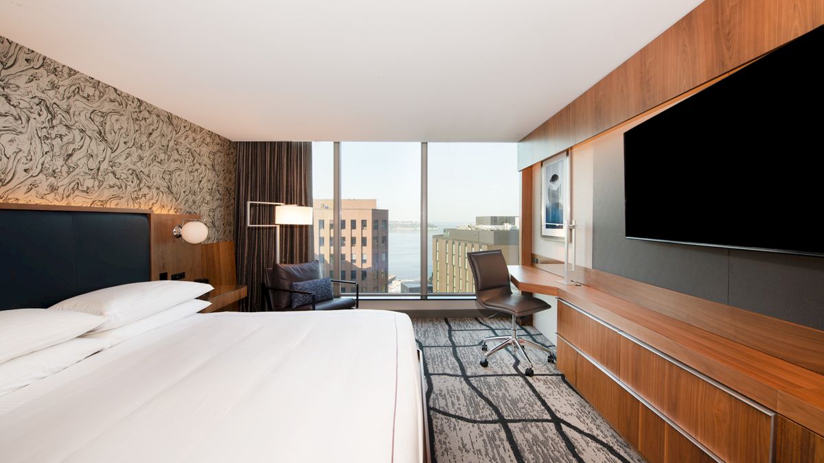 A modern hotel room features a bed, large TV, desk, and city view through floor-to-ceiling windows, with stylish decor and lighting.