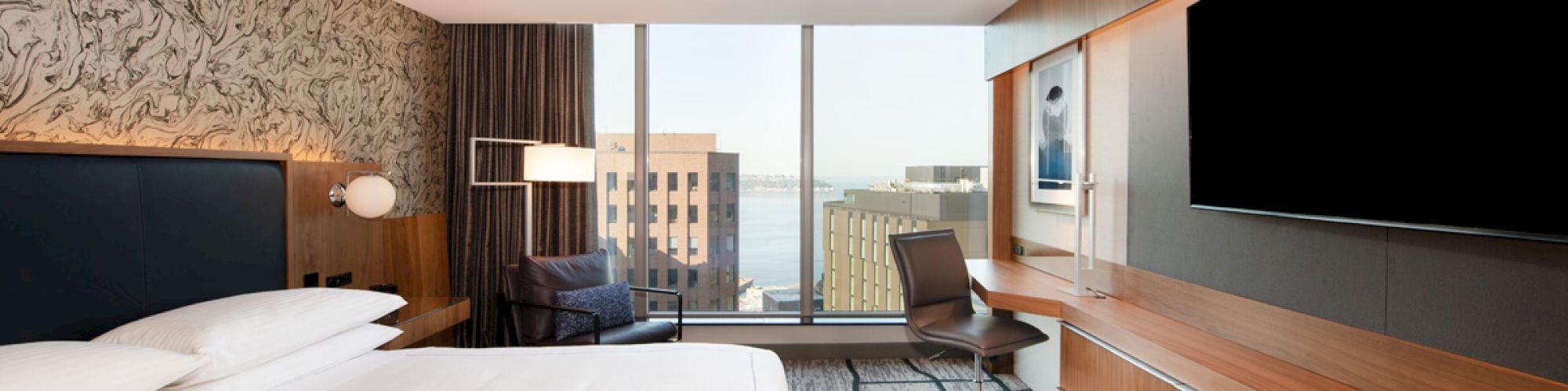 A modern hotel room features a bed, large TV, desk, and city view through floor-to-ceiling windows, with stylish decor and lighting.