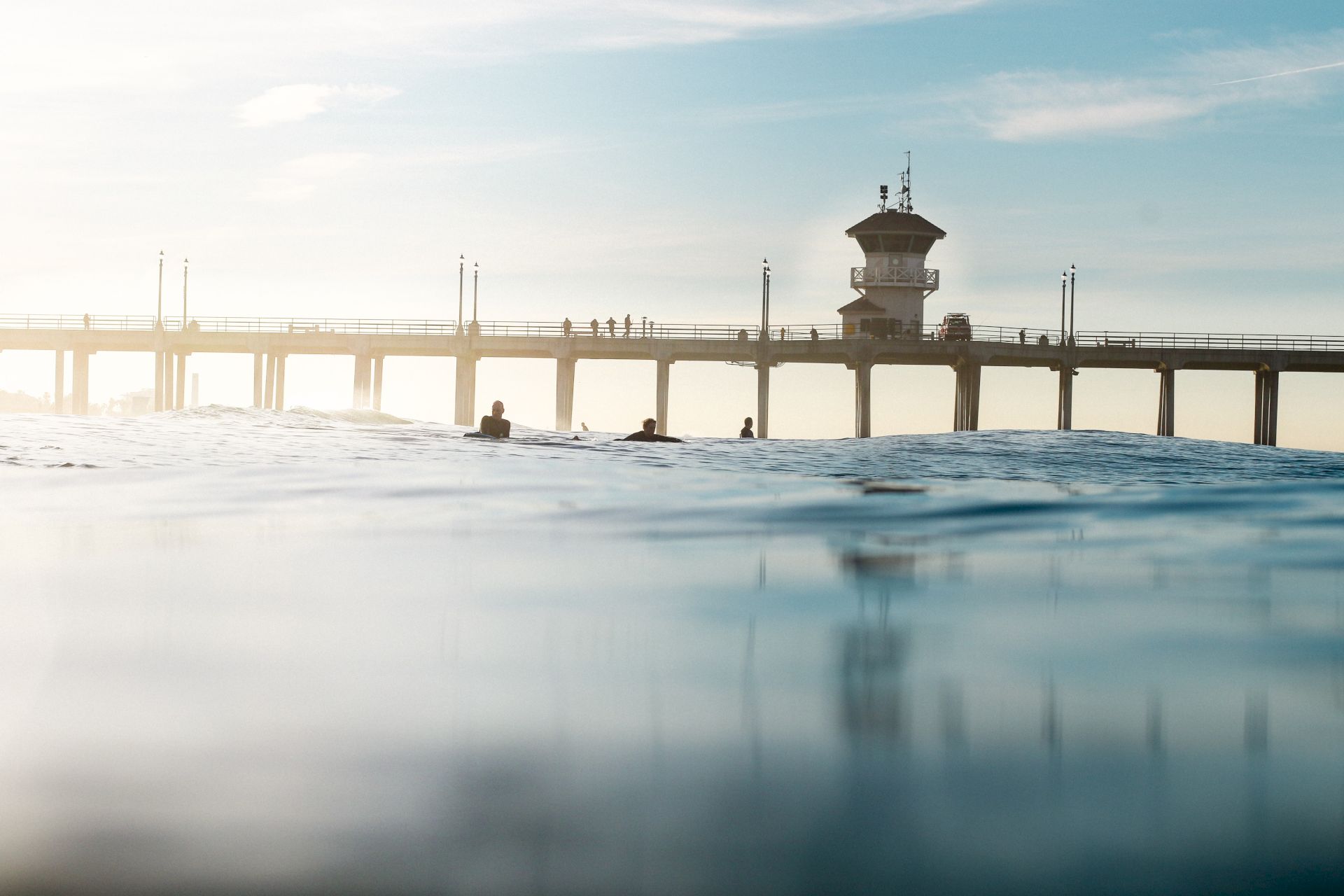 The image shows a beach pier with people walking, surfers in the water, and a partly cloudy sky in the background.